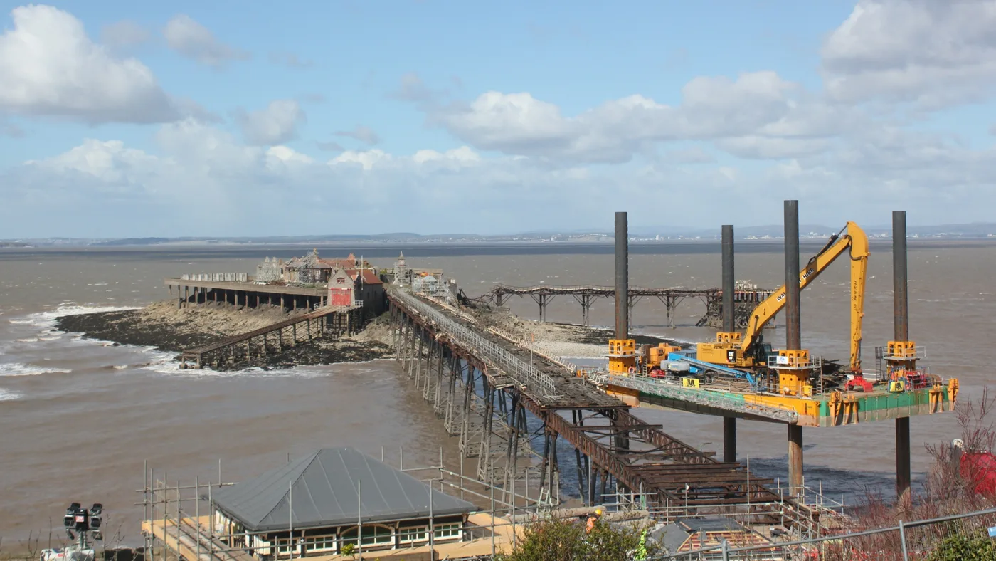 Restoration work on Birnbeck Pier, Weston-super-Mare, with the contractor's jack-up platform and excavator beside the stripped-back deck and Birnbeck Island beyond.
