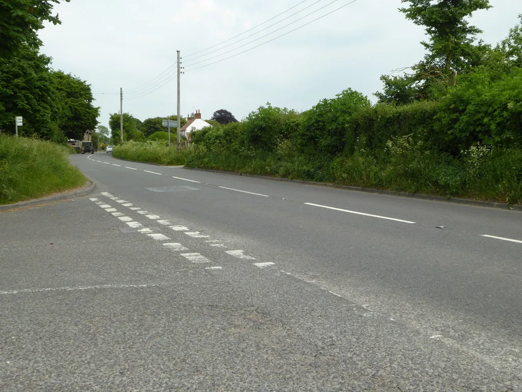 A two-lane country road lined with hedges and telegraph poles, curving away towards a house
