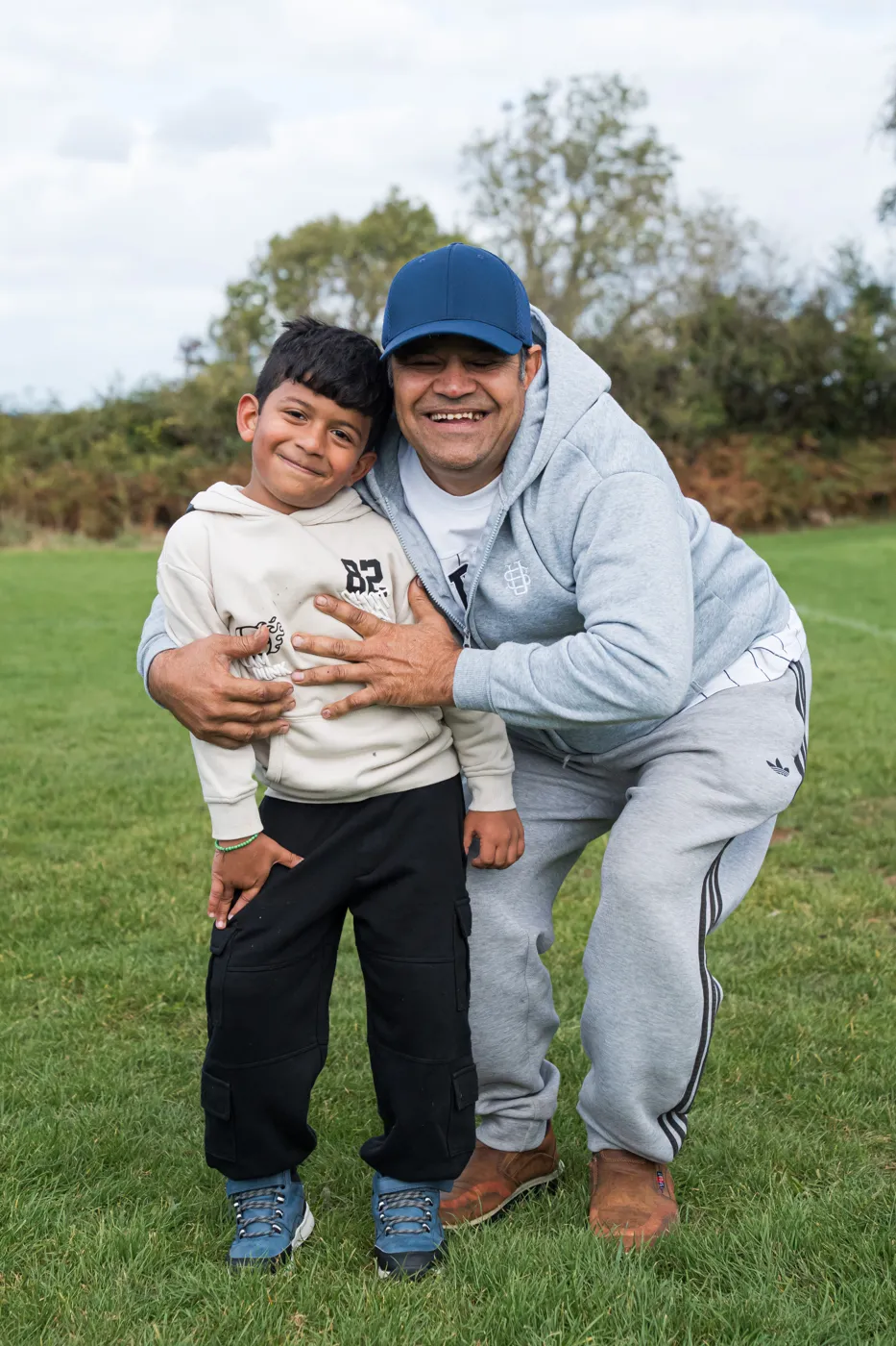 A smiling man in a blue cap and grey tracksuit crouching with his arm around a young boy on a grass field