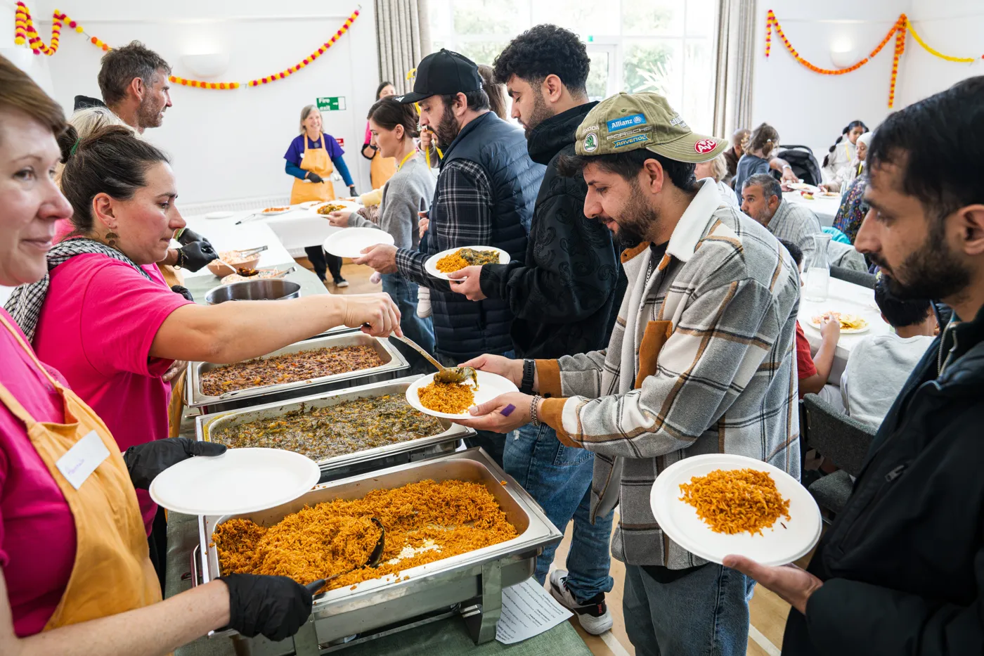 Volunteers in pink T-shirts and orange aprons serving rice and stew to a queue of guests holding plates