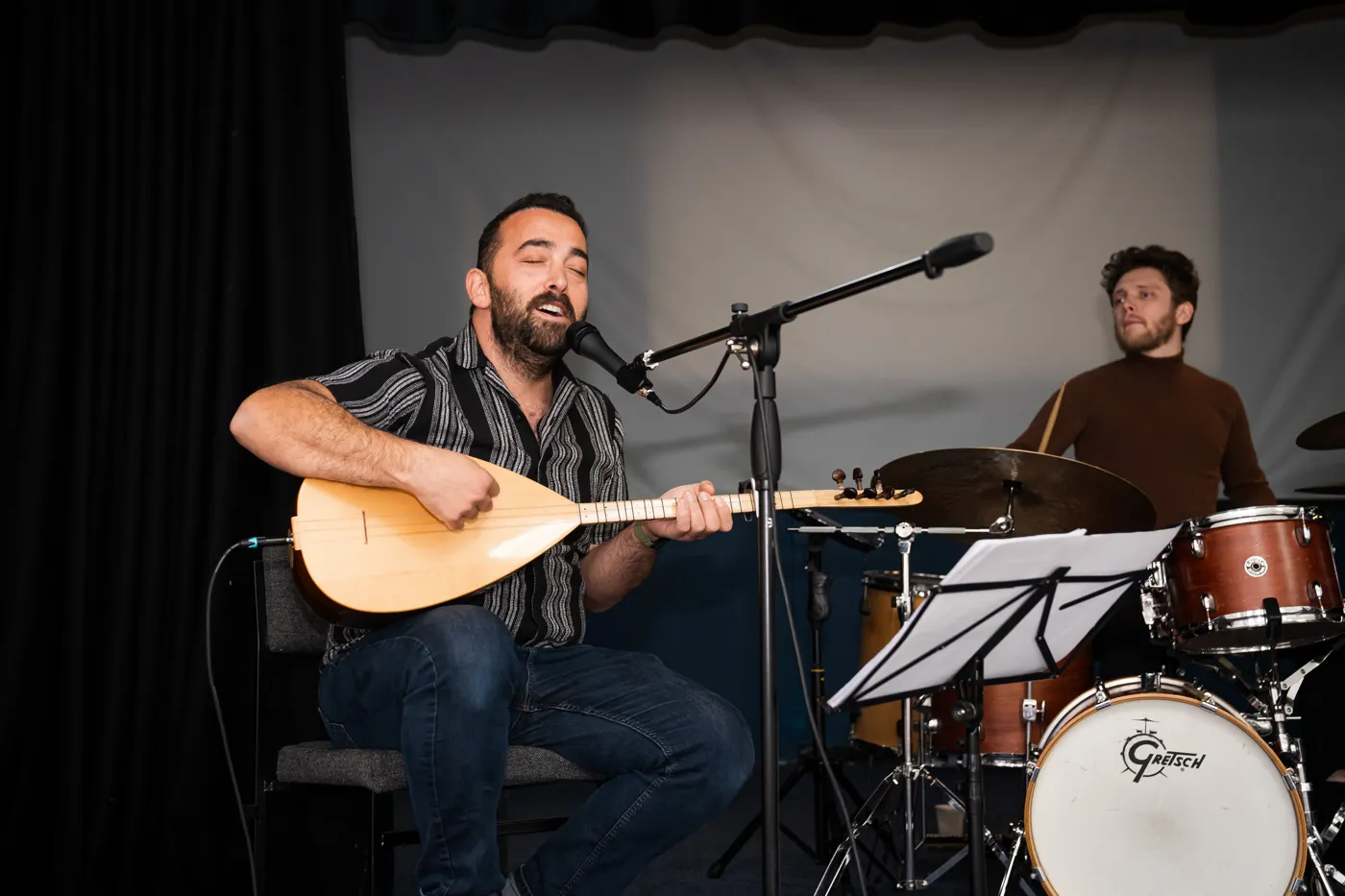 A musician singing and playing a long-necked stringed instrument at a microphone, with a drummer behind him