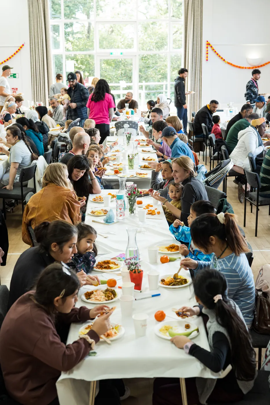 Adults and children eating at a long table covered with a white cloth in a hall decorated with orange and yellow garlands