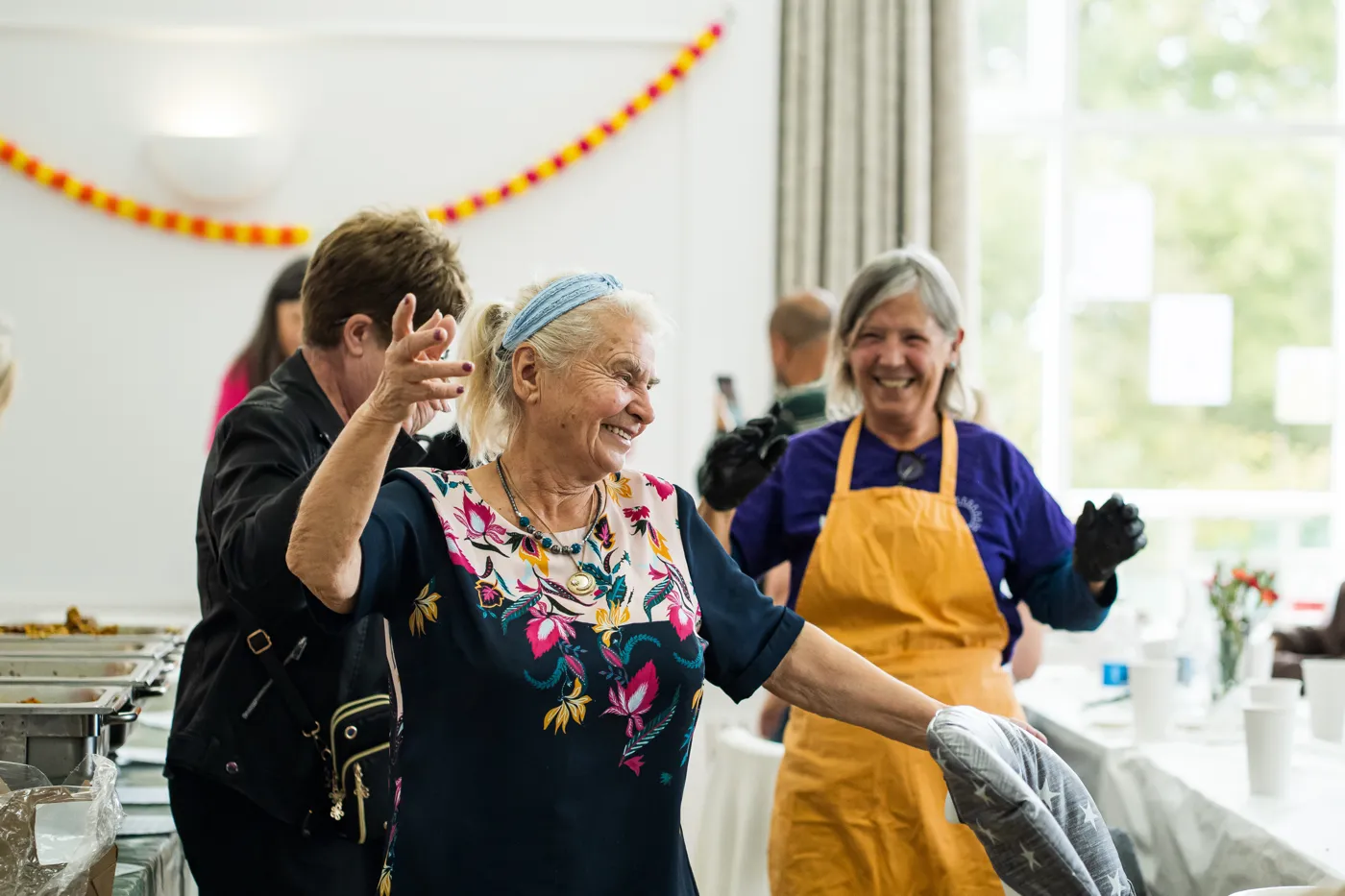 An older woman in a floral top dancing with her arms out, with a volunteer in an orange apron smiling behind her