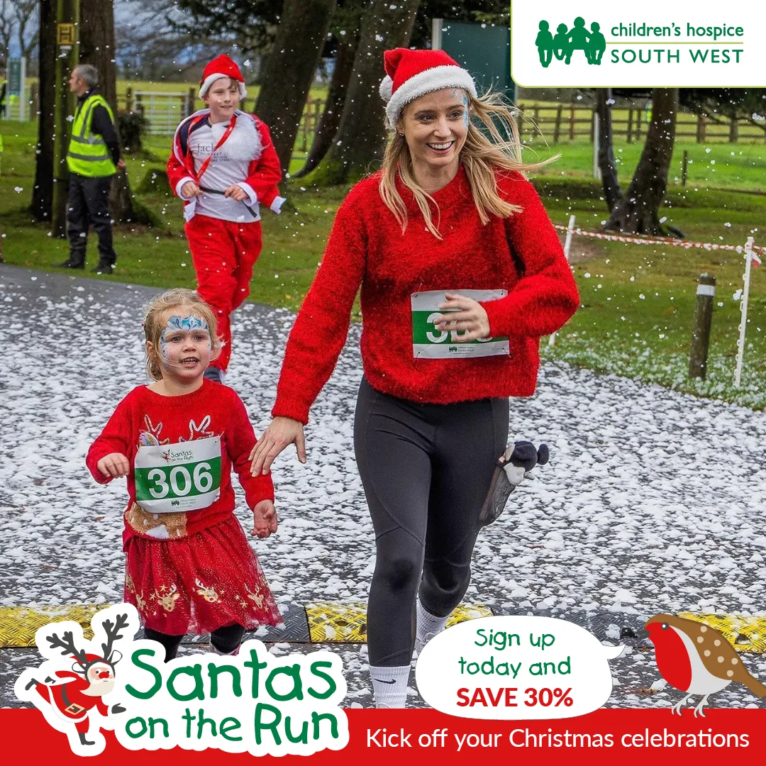 A woman in a red jumper and Santa hat runs hand in hand with a small girl in a reindeer dress wearing race number 306, on a path dusted with fake snow, under the Santas on the Run logo