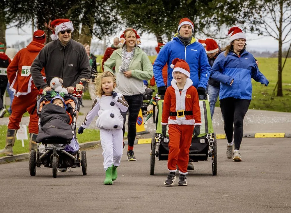 Families in Santa hats walking and jogging along a path, some pushing buggies, with a boy in a Santa suit and a girl dressed as a snowman