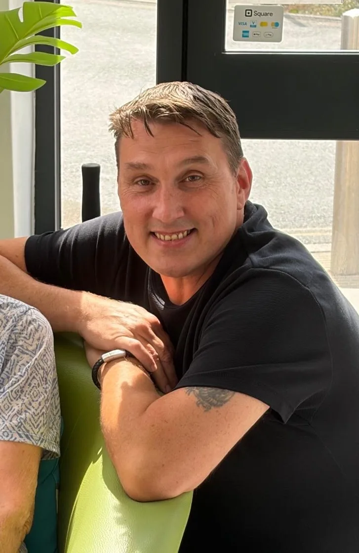 Andrew Cox of Toucan Law, smiling, leaning on the arm of a green sofa in a black T-shirt
